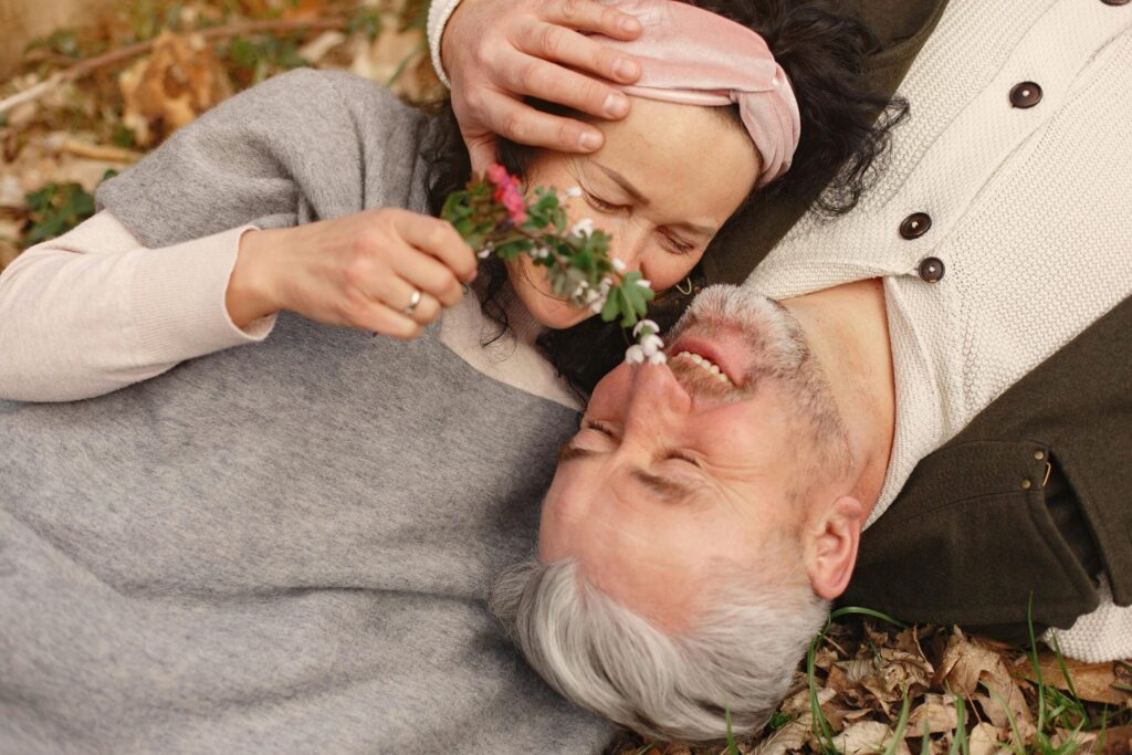 From above of cheerful senior wife wearing wide scarf and headband with flower bouquet in hand and happy elderly gray haired husband in warm clothes lying on ground with fallen leaves in park with closed eyes